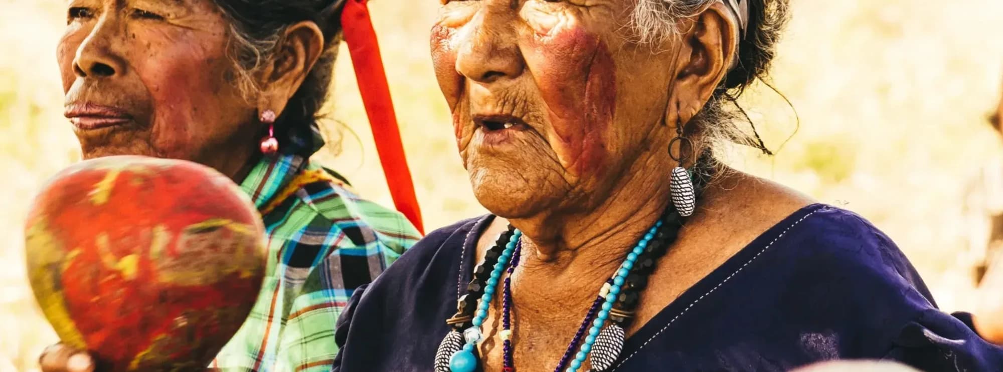 Two Indigenous women of Paraguay in traditional dress, one holding a painted gourd rattle